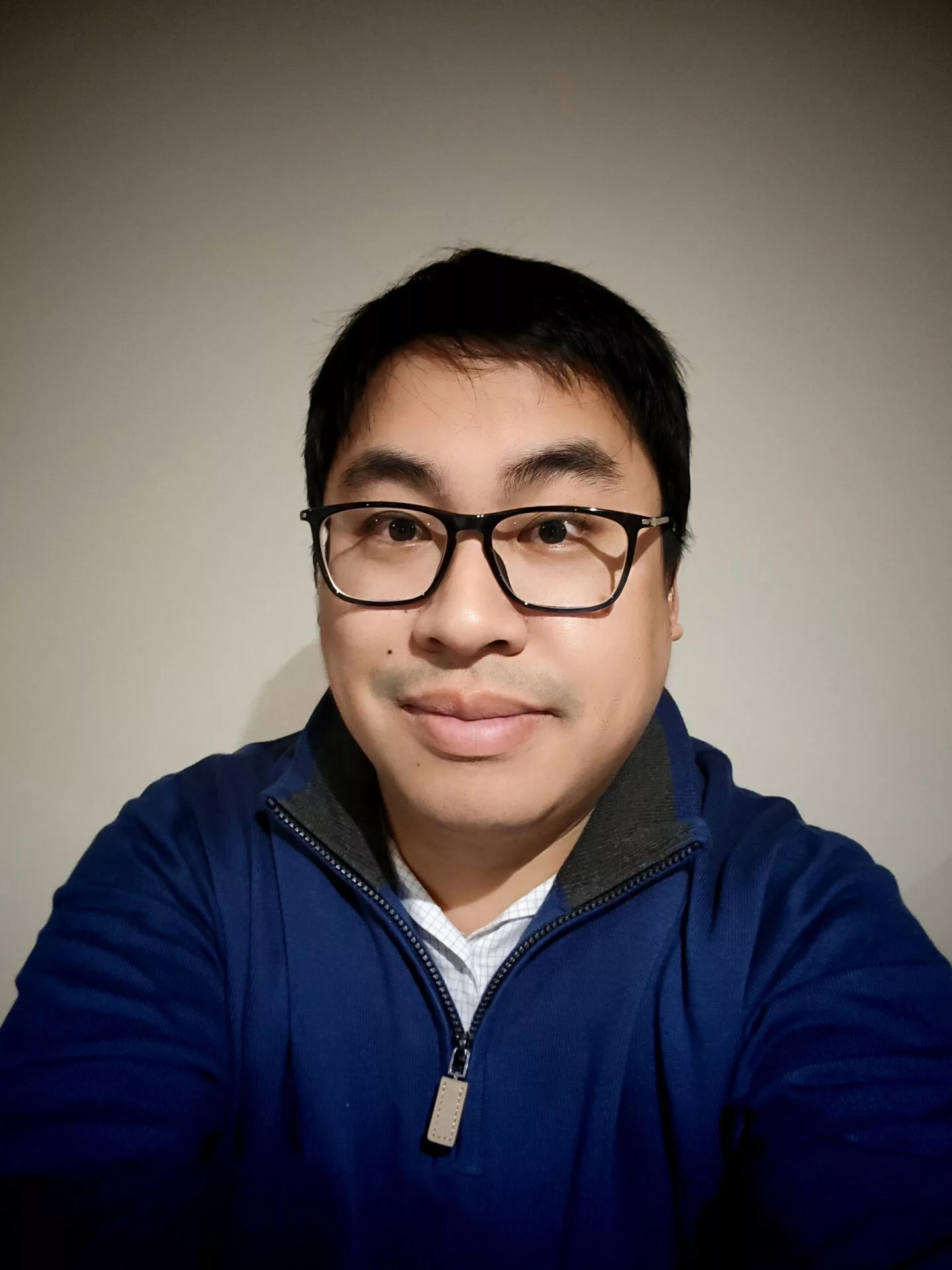InspireHM— A person with short black hair and glasses, wearing a blue zip-up sweater over a light shirt, is facing the camera and smiling slightly against a plain beige background.